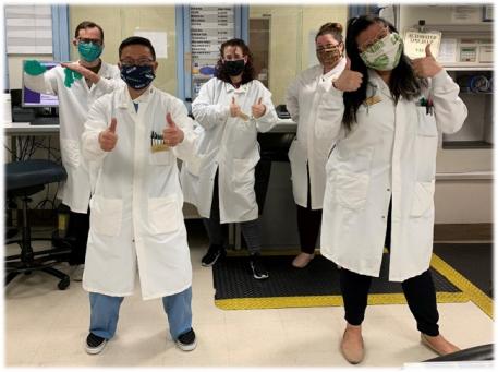 Five people in lab coats and face masks giving thumbs up to the camera